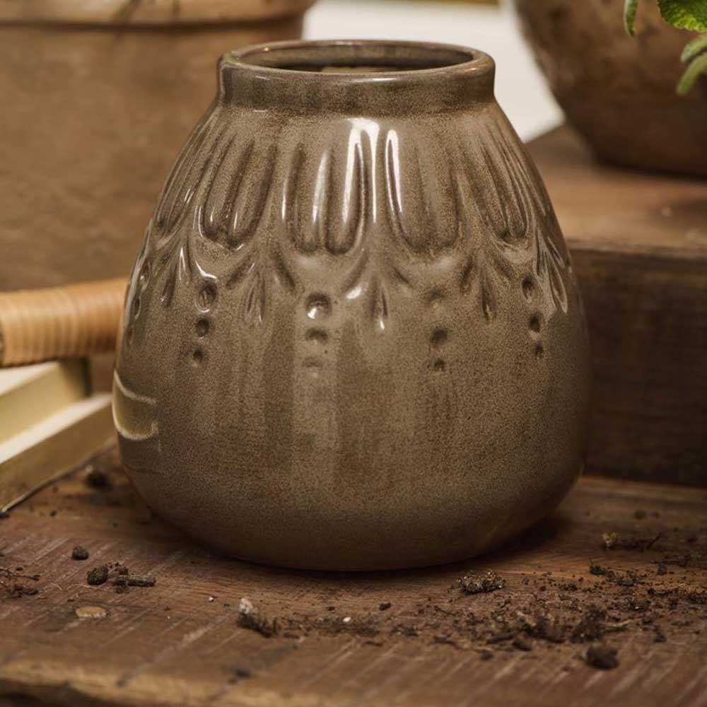 Eine kleine, braune Keramikvase von Ib Laursen mit einem strukturierten vertikalen Rillenmuster ruht auf einer Holzoberfläche, umgeben von Erde.