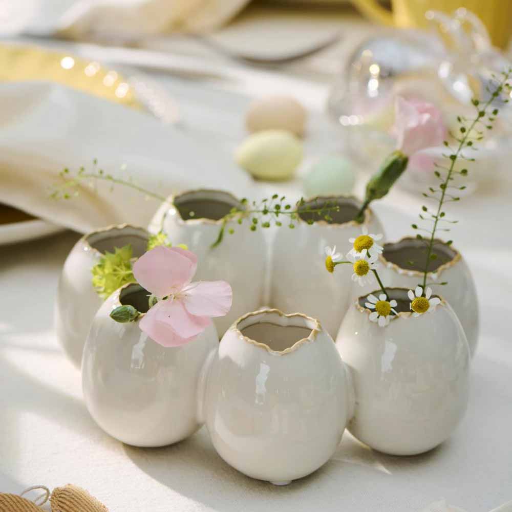Sechs Vasen von Ib Laursen, die wie zerbrochene Eierschalen gestaltet sind, enthalten kleine Blumen und Grünpflanzen und sind unter sanfter Beleuchtung auf einem Tisch arrangiert.