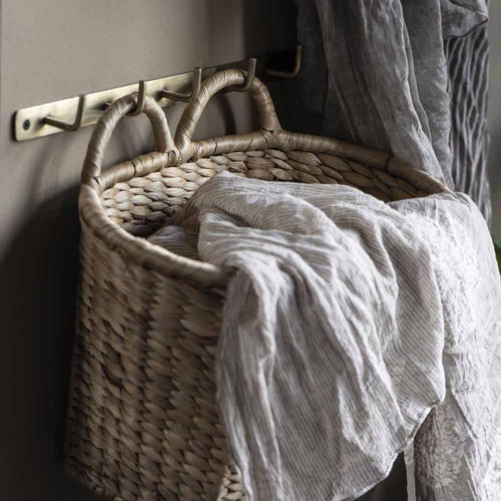 Ein viereckiger Wandkorb von Ib Laursen mit 2 Henkeln hängt an einem Messinghaken, darüber ist ein hellgrauer Stoff drapiert.