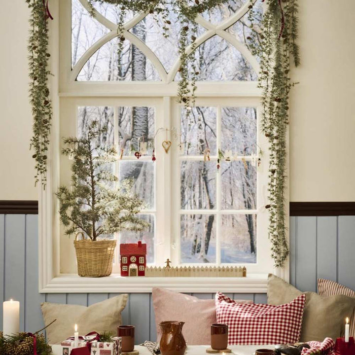 Ein gemütlicher Fensterplatz, winterlich dekoriert mit Ib Laursens Zederngirlande mit Zapfen, einem kleinen Tannenbaum in einem Korb, Kerzen, Tassen und festlichen Girlanden. Durch das Fenster sind schneebedeckte Bäume zu sehen.