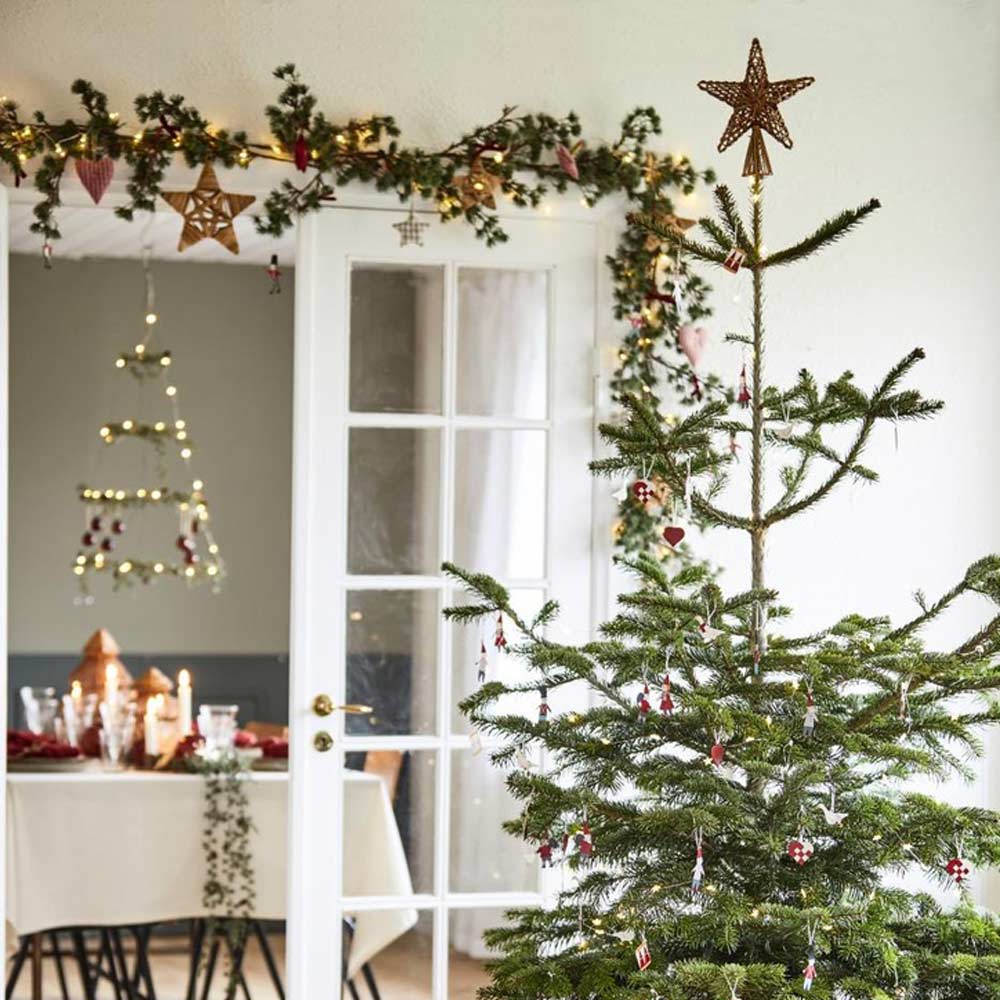 In einem Wohnzimmer steht ein geschmückter Weihnachtsbaum mit einem Stern an der Spitze und eine Ib Laursen Zederngirlande mit Zapfen ist festlich um eine Tür geschmückt. In einem angrenzenden Raum ist ein Esstisch aufgestellt.