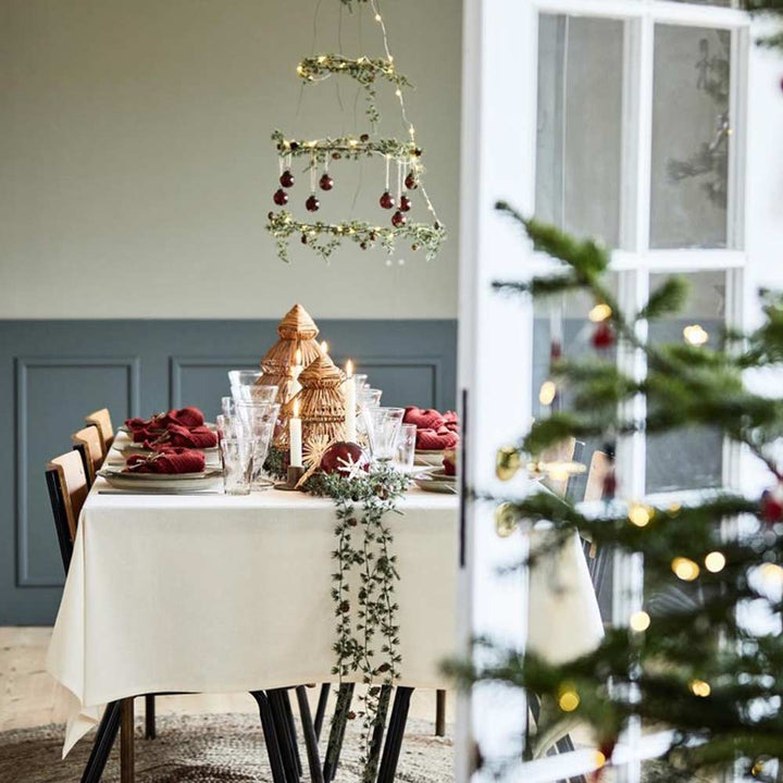 Festlich gedeckter Esstisch mit roten Servietten, Glaswaren und Dekoelementen. Darüber hängt ein Kronleuchter, geschmückt mit der Ib Laursen Zederngirlande mit Zapfen und Ornamenten. Im Vordergrund ist teilweise ein Weihnachtsbaum mit Lichtern zu sehen.