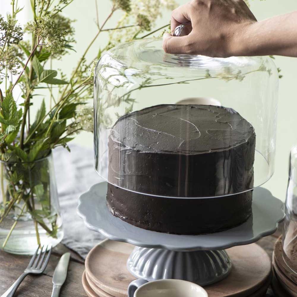 Eine Hand hebt eine Ib Laursen - Glasglocke für Tortenplatte von einem Schokoladenkuchen auf einem Kuchenständer, daneben eine Vase mit Blumen und Utensilien.