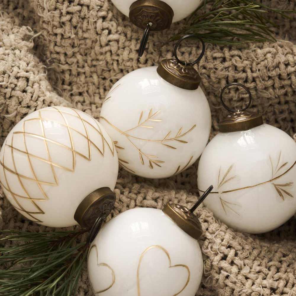 Ein Set aus vier weißen Ornamenten von Ib Laursen mit aufwendigen Goldgravuren ruht auf einem strukturierten beigen Tuch, elegant ergänzt durch grüne Kiefernnadeln.