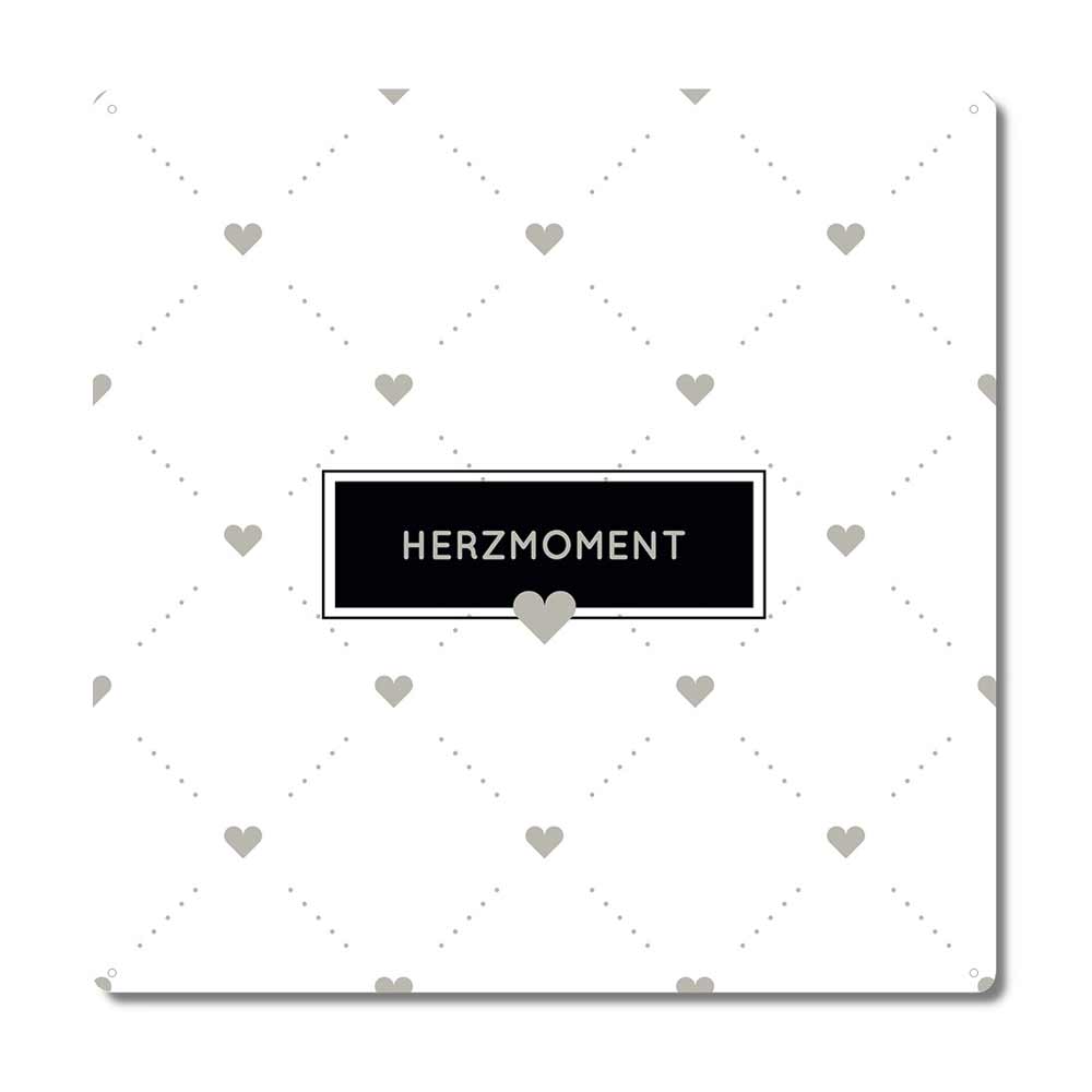 Ein weißer Hintergrund mit einem Muster aus kleinen grauen Herzen, die diagonal angeordnet sind. In der Mitte ein schwarzes rechteckiges Etikett mit dem Wort „Interluxe Metallschild – Herzmoment“ und darunter ein graues Herz.