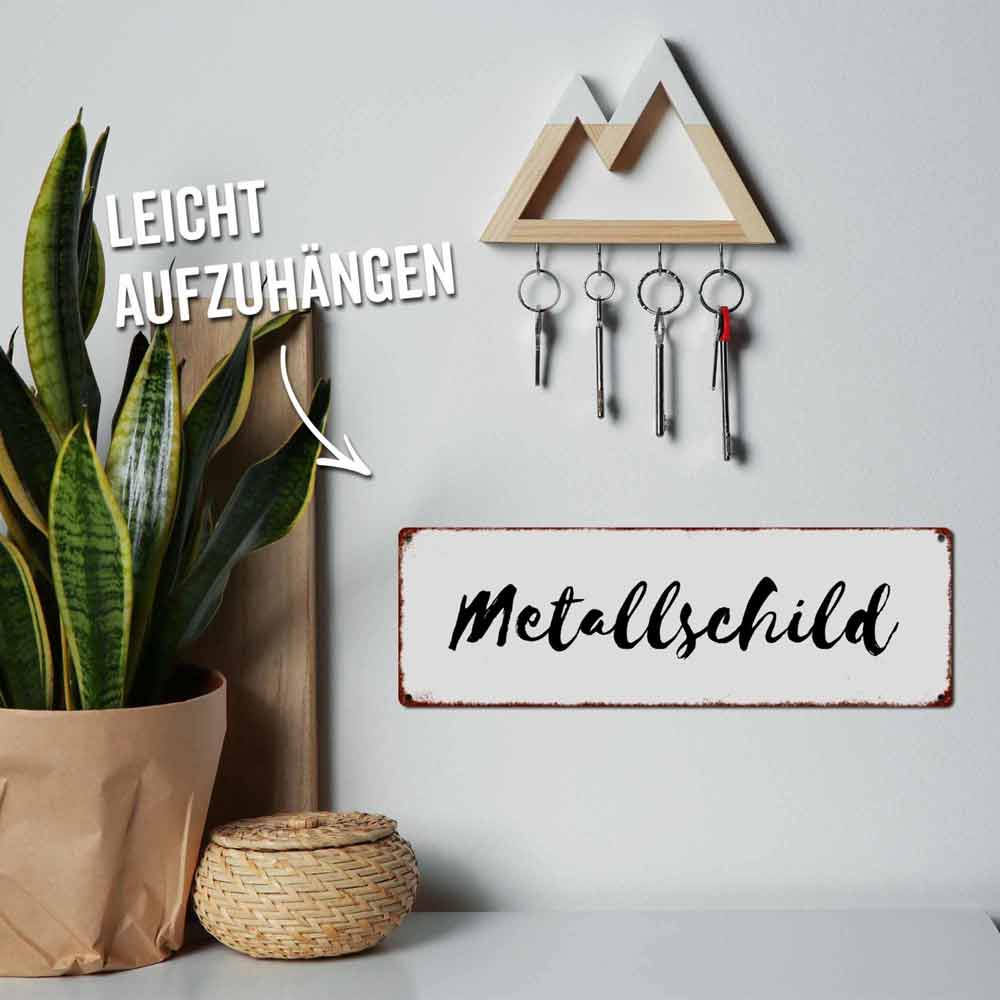 Unter einem dekorativen Regal hängen eine kleine Pflanze in einer Papiertüte, ein Weidenkorb und ein wandmontierter Schlüsselhalter mit fünf Schlüsseln. An der Wand hängt ein Schild mit der Aufschrift „Interluxe Metallschild – Liebes Wochenende nicht so schüchtern“. Text lautet „Leicht aufzuhängen.