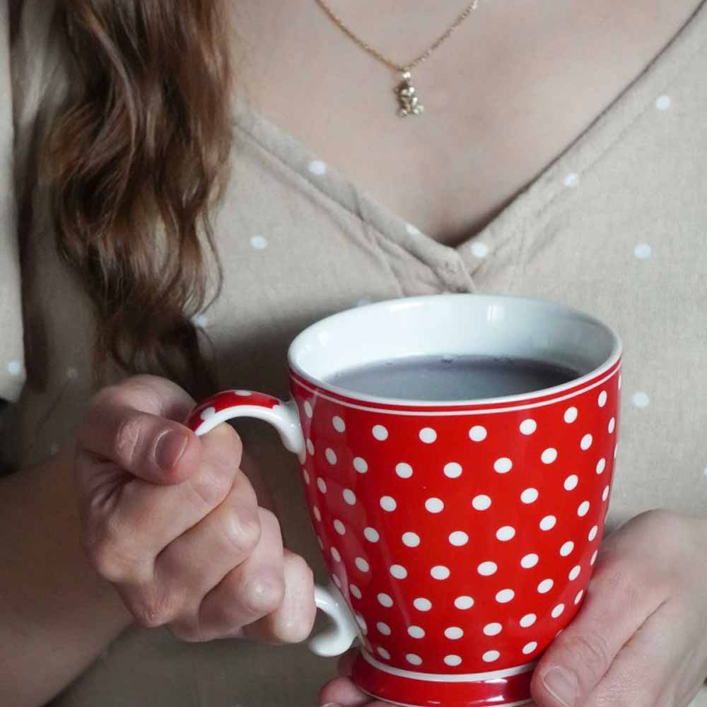 Eine Person hält die Isabelle Rose – Tasse Keramik mit Punkten Rot, gefüllt mit einer dunklen Flüssigkeit, und trägt ein beiges Oberteil mit kleinen weißen Punkten.