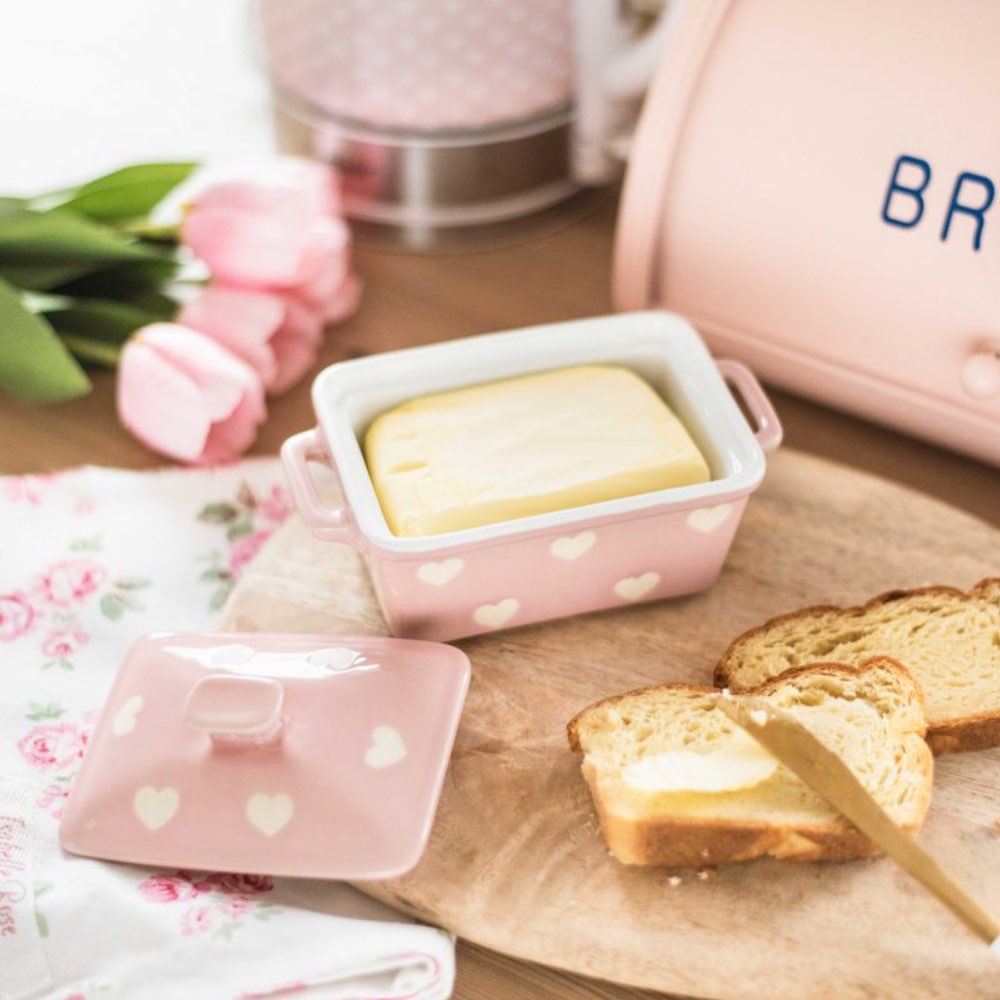 Die Isabelle Rose - Butterdose mit Herzen, eine rosafarbene Butterdose mit Herzmuster, hält Butter neben zwei Scheiben Toast auf einem Holzbrett, begleitet von rosafarbenen Tulpen und einer Blumenserviette.