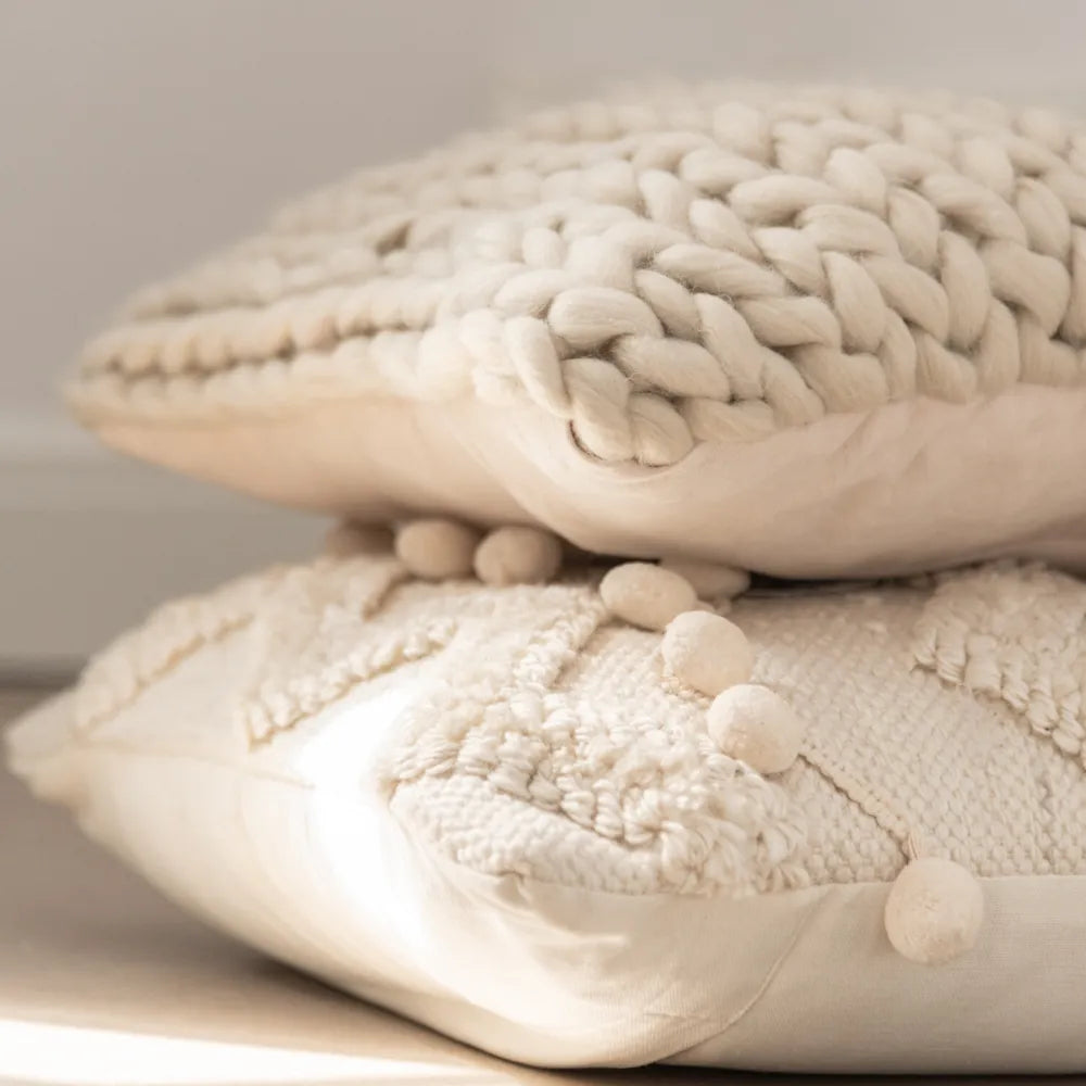 Helle Kissen übereinander, oben große Strickmaschen, darunter glatte Stoffflächen und kleine Bommeln