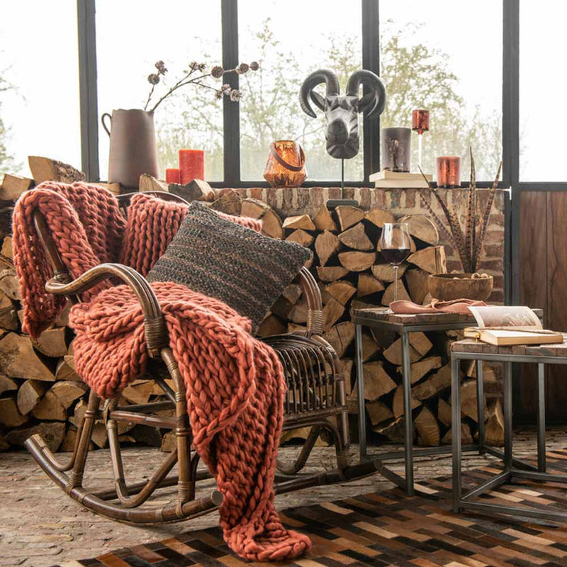 Rattansessel mit rostrotem Strick und grauem Kissen vor gestapeltem Holz