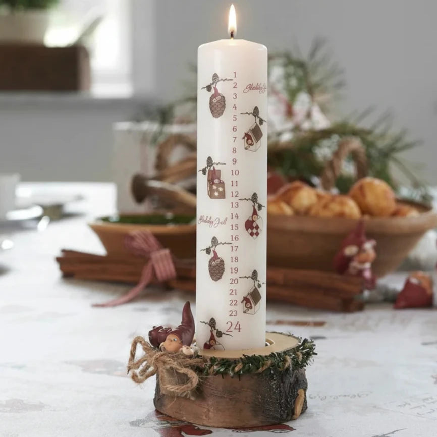 Die Klarborg Denmark - Adventskerze 1-24 Nisserik White ist eine brennende Adventskerze, nummeriert von 1 bis 24, auf einem hölzernen Sockel mit verschwommener Weihnachtsdekoration und Essen im Hintergrund.