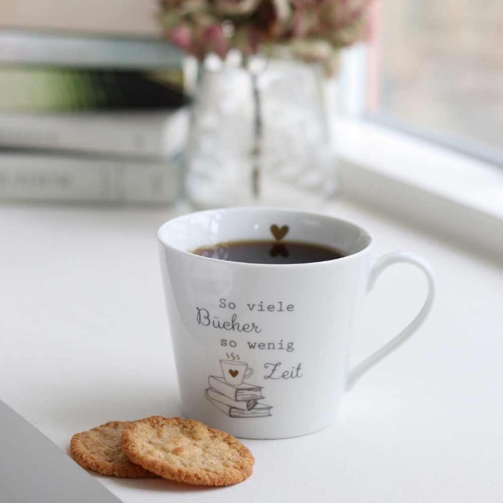 Eine Krasilnikoff - Happy Cup Lesen macht glücklich-Tasse mit deutschem Text steht neben zwei Keksen auf einem Tisch, im Hintergrund sind mehrere Bücher gestapelt.