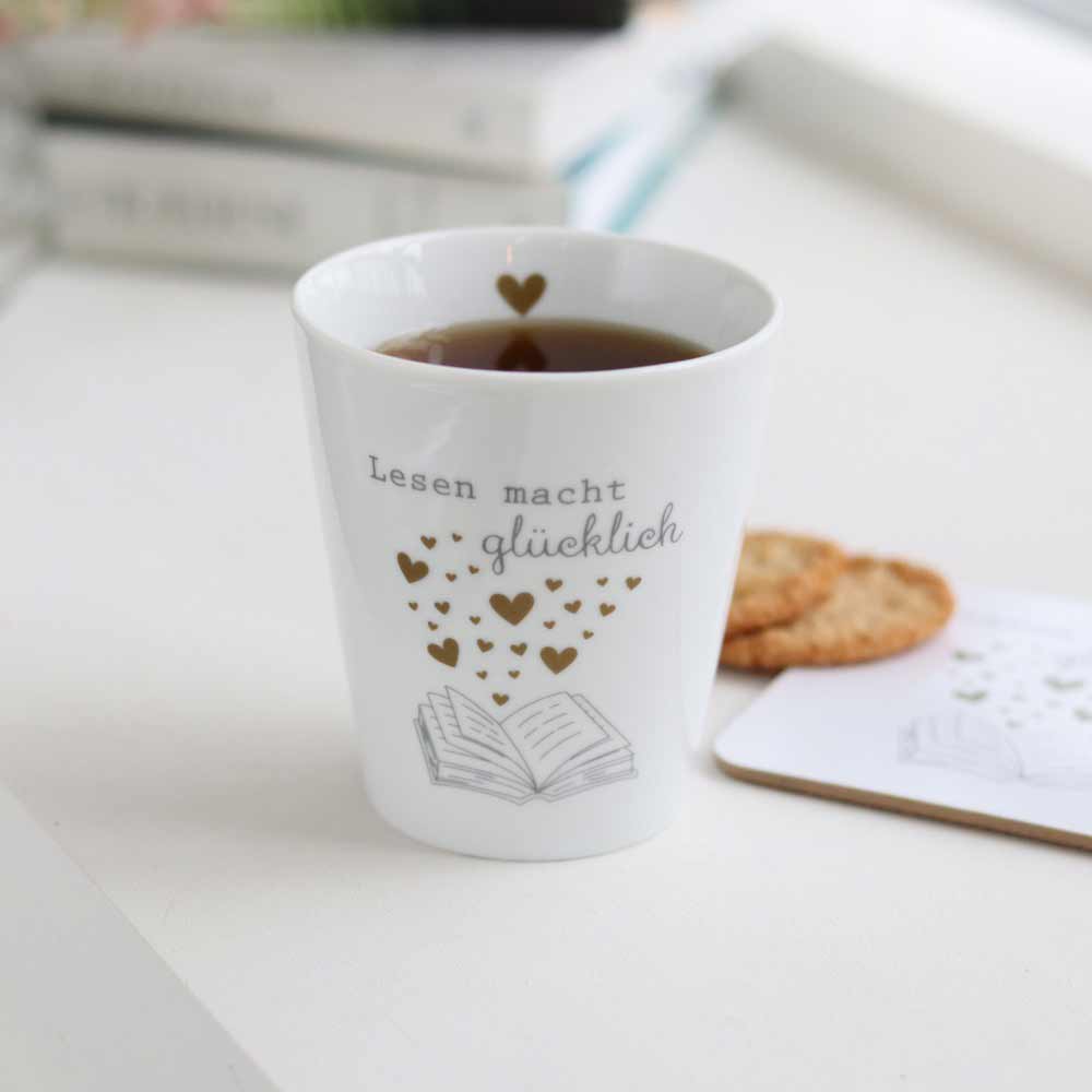 Eine Krasilnikoff - Happy Mug „Lesen macht glücklich“ mit goldenen Herzen und dem deutschen Text „Lesen macht glücklich“ neben einem Keks und einer Illustration eines aufgeschlagenen Buches.