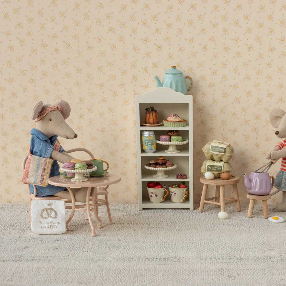 Das Maileg Café-Set Tisch & Bank besteht aus zwei ausgestopften Mäusen, die in einem Puppenhaus Tee trinken, mit einem Schrank mit Gebäck und einer Teekanne. Eine Maus trägt einen Rucksack, während kleine Hocker und eine Einkaufstasche in der Nähe stehen.