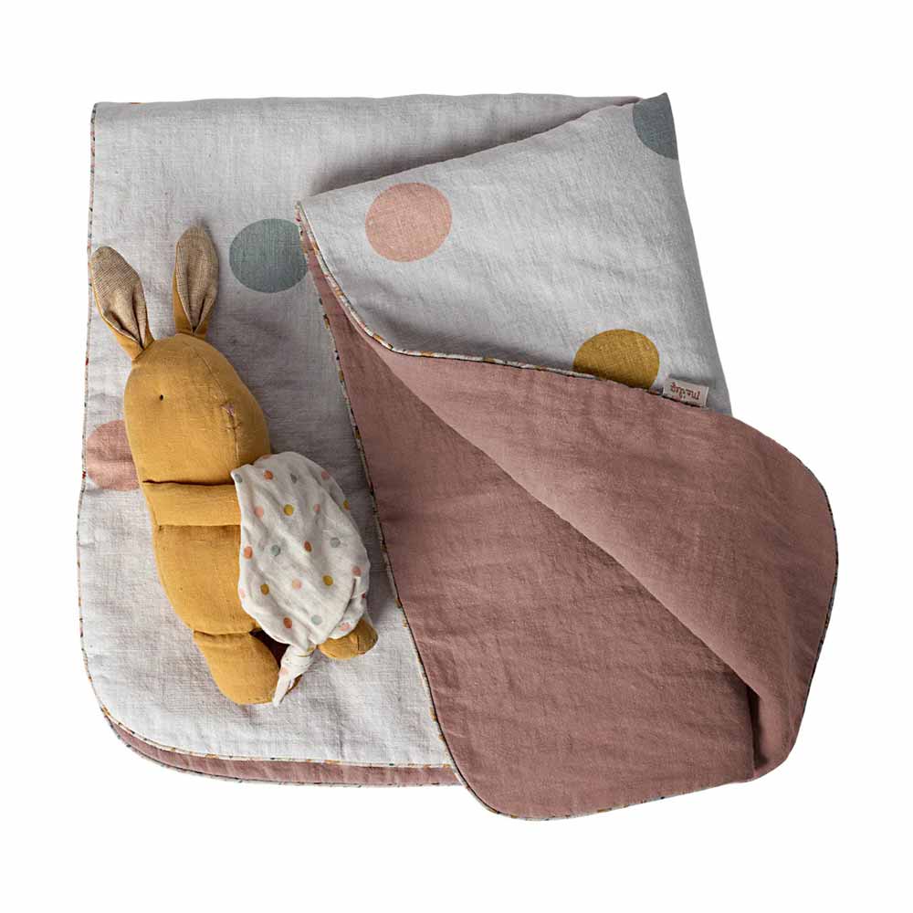 Ein Stoffhase, der ein Tuch hält, sitzt auf einer gefalteten Maileg-Decke „Baby Rose mit Punkten“ mit einem gepunkteten und einfarbigen Design in Beige-, Rosa- und Brauntönen.