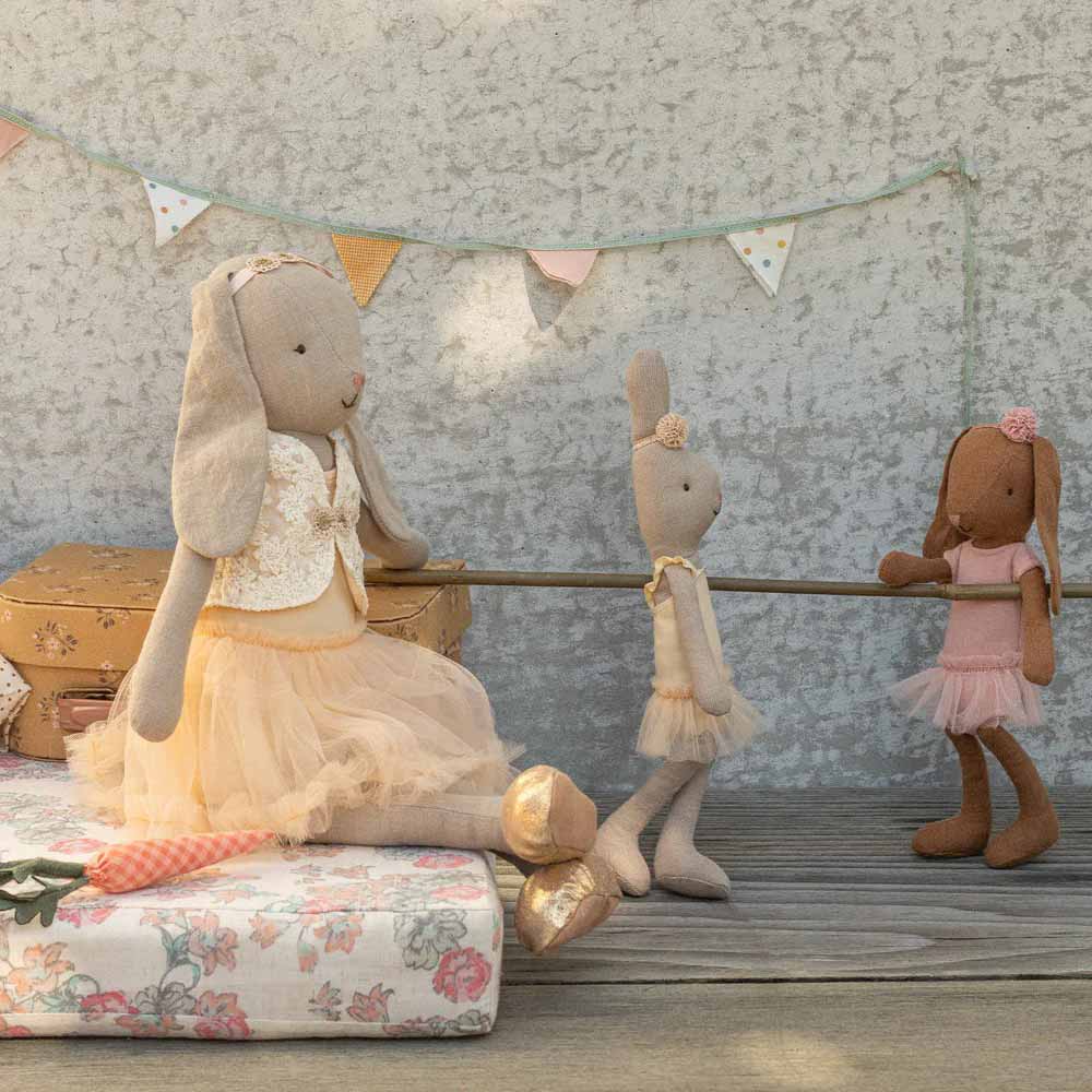 Drei Maileg Hase Bunny Ballerina Größe 1 Puppen in pastellfarbenen Kostümen scheinen gemeinsam im Freien zu spielen, wobei alte Koffer und Blumenwimpel den Hintergrund schmücken.