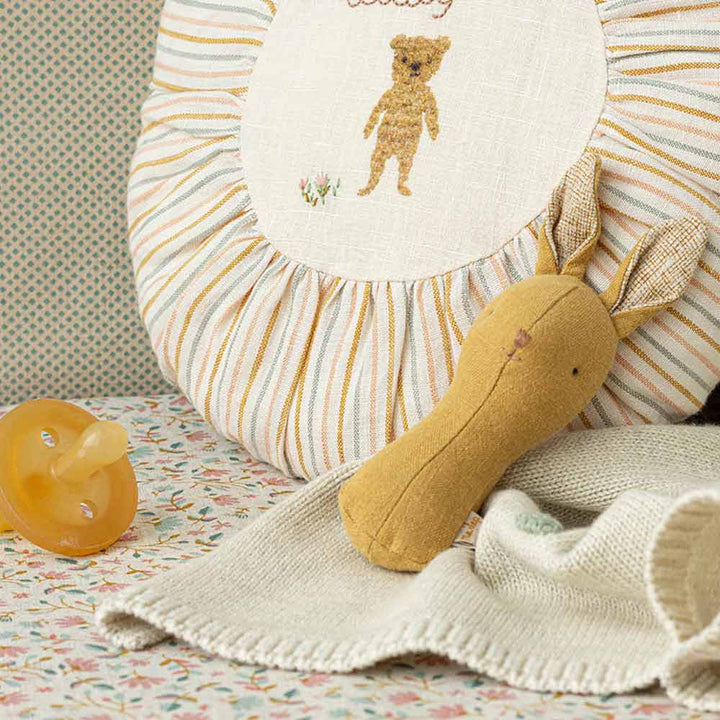 Ein Maileg - Hase Rassel Lullaby Friends-Häschenspielzeug, eine beige Strickdecke und ein gelber Schnuller liegen auf einer gemusterten Oberfläche vor einem runden Kissen mit einem darauf gestickten Bären.