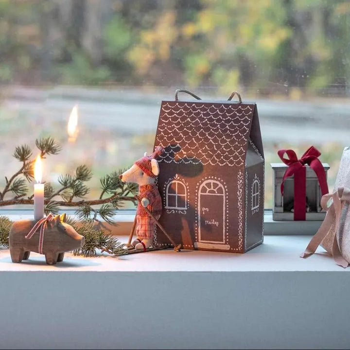 Die Maileg Lebkuchenhaus Tasche Geschenkverpackung steht auf einer Fensterbank neben einer Kerze, einer Rentierfigur, einem Spielzeugtier, einer Geschenkverpackung und Grünzeug.