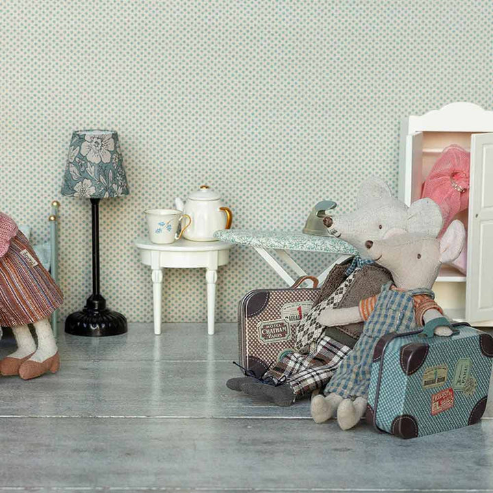 Zwei Plüschmäuse sitzen mit Koffern in einem Puppenhauszimmer, das ein Teeservice, ein Bügelbrett, eine Lampe und einen Kleiderschrank enthält. Links sind teilweise die Beine einer weiteren Maus zu sehen. Das Produkt ist Maileg - Maus Big Brother Overall und Hemd im Koffer.
