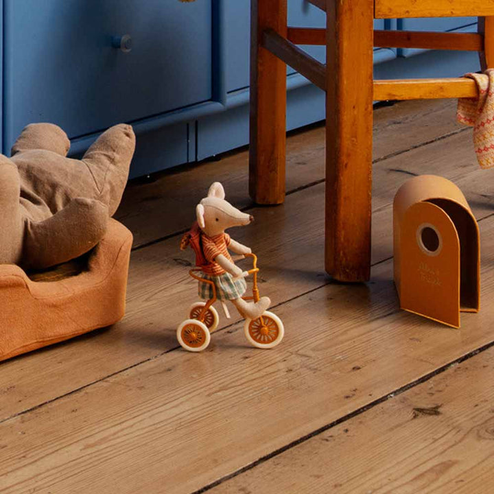 Eine Maileg-Maus mit Hut fährt auf einem kleinen Dreirad über einen Holzboden in der Nähe eines Plüschsessels und eines Spielzeug-Mauselochs.
