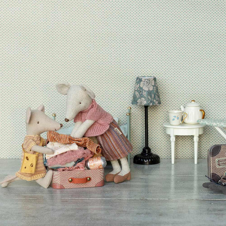 Zwei Spielzeugmäuse packen einen kleinen Koffer vor einer gemusterten Wand; eine Maus steht, während die andere kniet, neben einer Lampe, einem Teekannen-Set, einem Bügelbrett und „Maleg – Maus Big Sister“, gesttricktes Kleid und Tasche im Koffer.