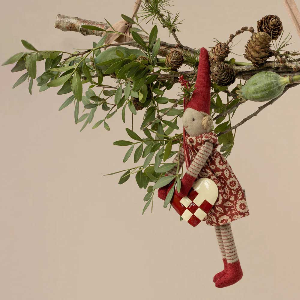 Das Maileg - Metallornament Herz besteht aus einer Elfenpuppe aus Stoff in einem roten Outfit, die ein kariertes Herzornament hält, das mit grünen Blättern und Tannenzapfen geschmückt ist.