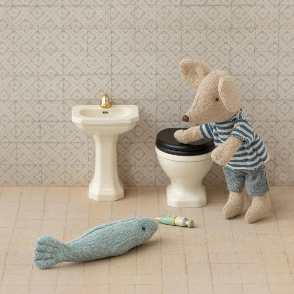 Eine Maileg Miniatur Waschbecken Maus in einem gestreiften Hemd steht neben einem Spielzeug-Waschbecken und einer Toilette, gegenüber einem Spielzeugfisch und einer kleinen Röhre auf dem gefliesten Boden.
