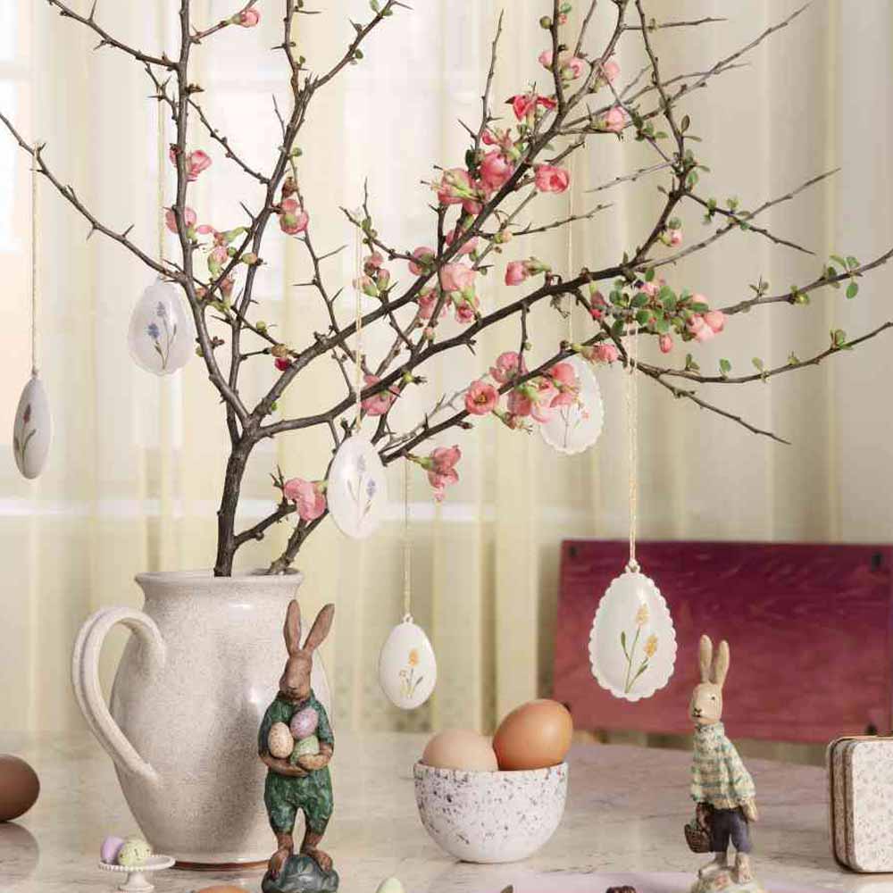 Eine dekorative Präsentation zum Thema Ostern mit einer Vase, in der ein blühender Zweig steht, geschmückt mit Maileg-Ostereier-Metallanhänger-2er-Set-Ornamenten, flankiert von Hasen- und Eierfiguren auf einem Tisch.