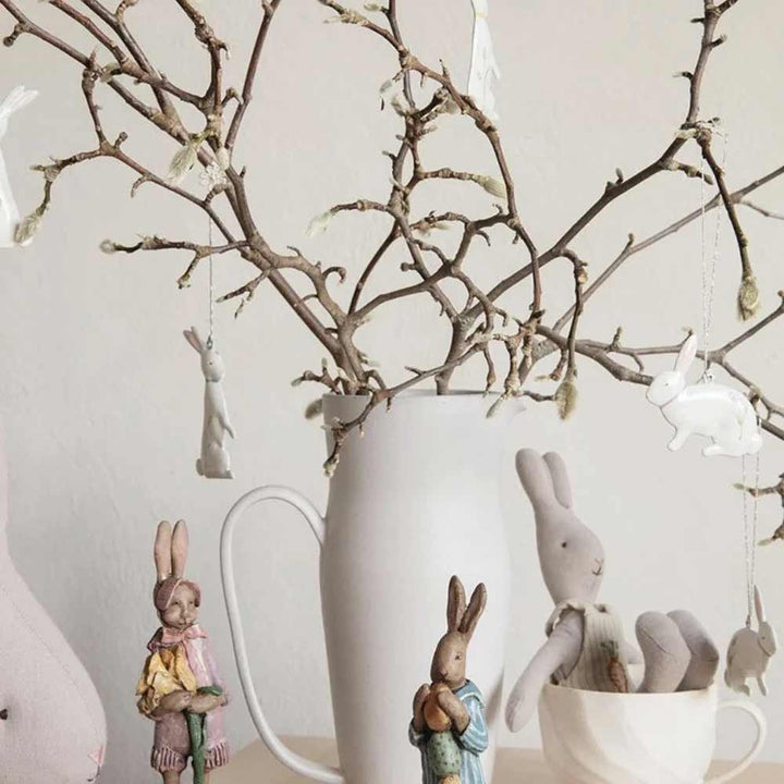 Eine Ausstellung von Maileg – Osterhasen-Metallanhänger, 5er-Set in Box mit Blumenmuster, arrangiert um und hängend an kahlen Zweigen in einer weißen Vase.