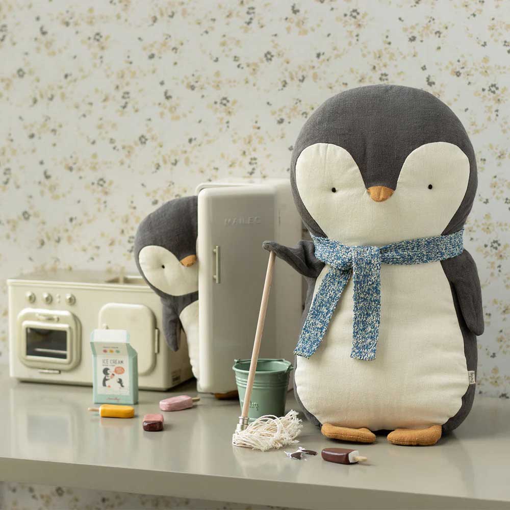 Zwei Maileg-Pinguine in einer Küchenumgebung, einer hält einen Wischmopp. Auf der Theke stehen ein Spielzeugherd, ein Kühlschrank, ein Eisschild und Reinigungsutensilien.