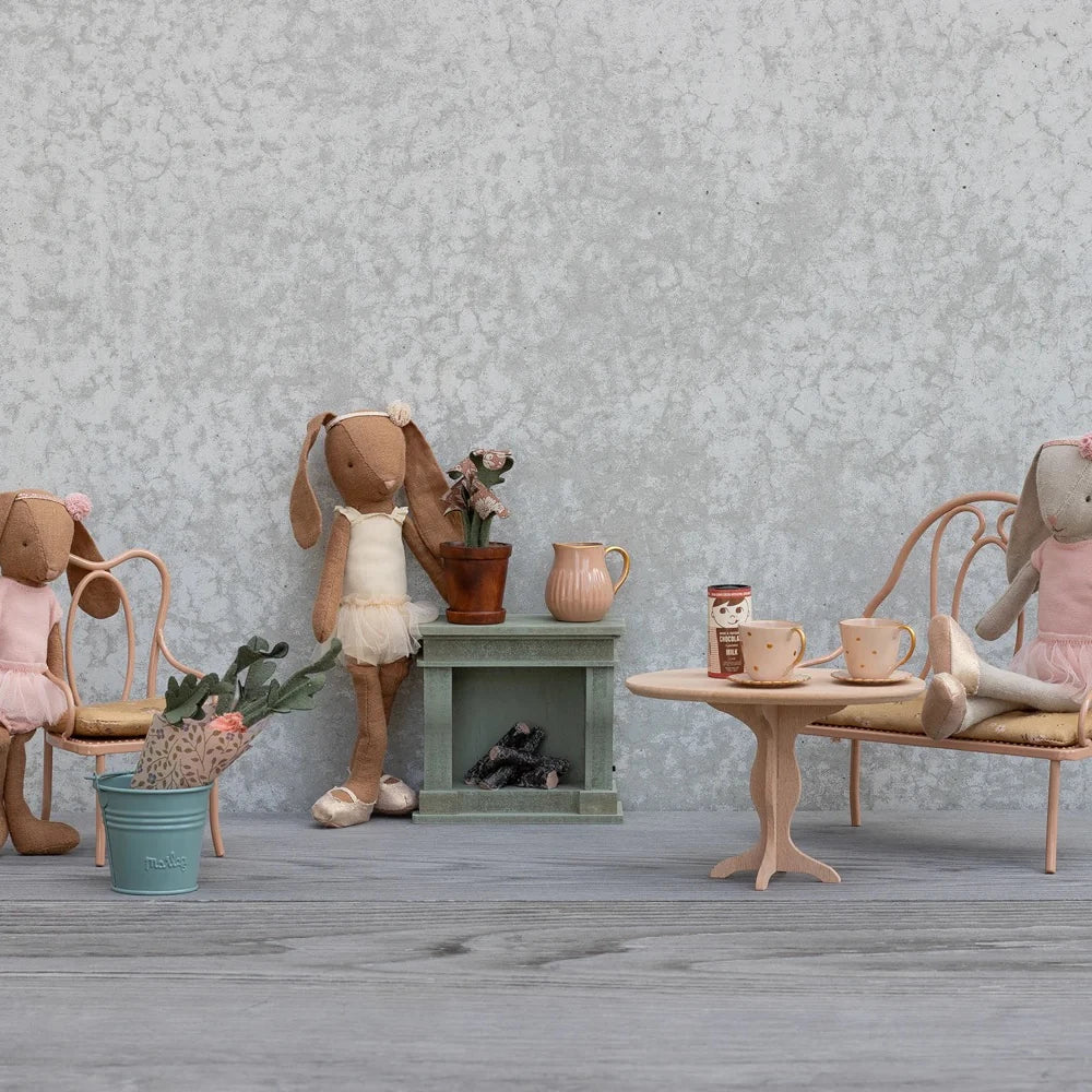 Drei ausgestopfte Hasenpuppen in rosafarbenen Outfits sitzen um Maileg - Puppenmöbel Vintage Stuhl Dunkelrosa und andere Miniaturmöbel, darunter ein Tisch mit Tassen, ein Regal, Blumen und Stühle, die alle vor einem strukturierten grauen Hintergrund stehen.