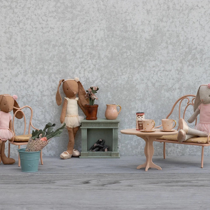 Drei ausgestopfte Hasenpuppen in rosafarbenen Outfits sitzen um Maileg - Puppenmöbel Vintage Stuhl Dunkelrosa und andere Miniaturmöbel, darunter ein Tisch mit Tassen, ein Regal, Blumen und Stühle, die alle vor einem strukturierten grauen Hintergrund stehen.