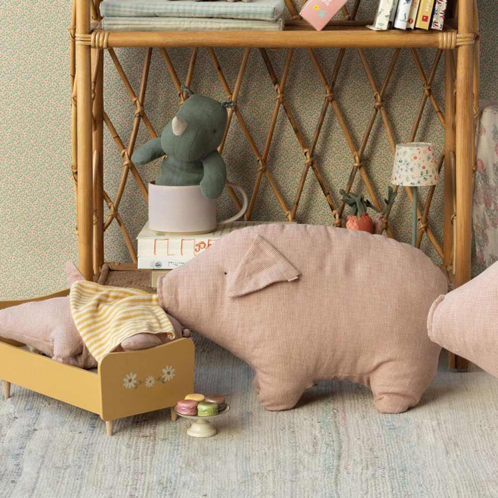 Eine Kinderzimmerszene mit einem Regal voller Spielbücher und einem weichen Teddybären. Es gibt ein Maileg - Schwein Polly Pork und ein Spielbett, in dem eine kleine Stofffigur liegt. Verschiedene kleine Gegenstände liegen verstreut herum.