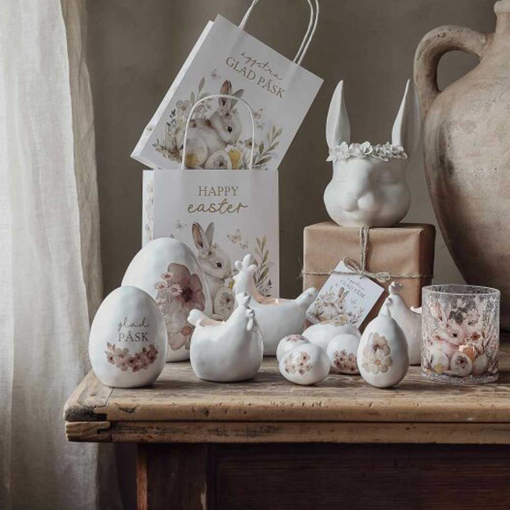„Deko Ei mit Blume“ von Majas Cottage sorgt zusammen mit Keramikeiern, Hasen und Hühnern mit floralen Motiven für eine festliche Osterszene auf einem Holztisch, im Hintergrund mit Geschenktüten und Geschenken geschmückt.