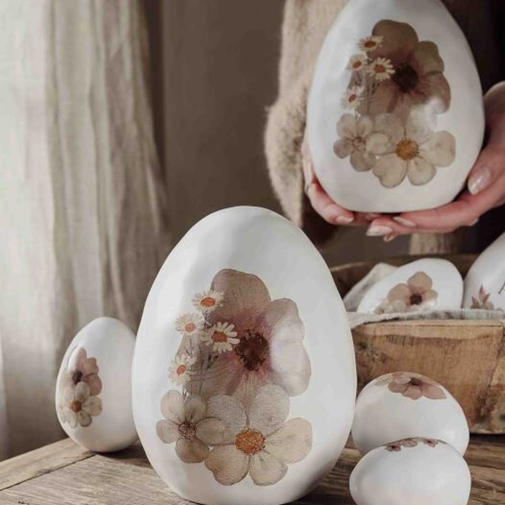 Hände halten ein Majas Cottage – Deko-Ei mit Blume, das zwischen anderen Deko-Eiern mit gepressten Blumenmotiven auf einer Holzoberfläche präsentiert wird.