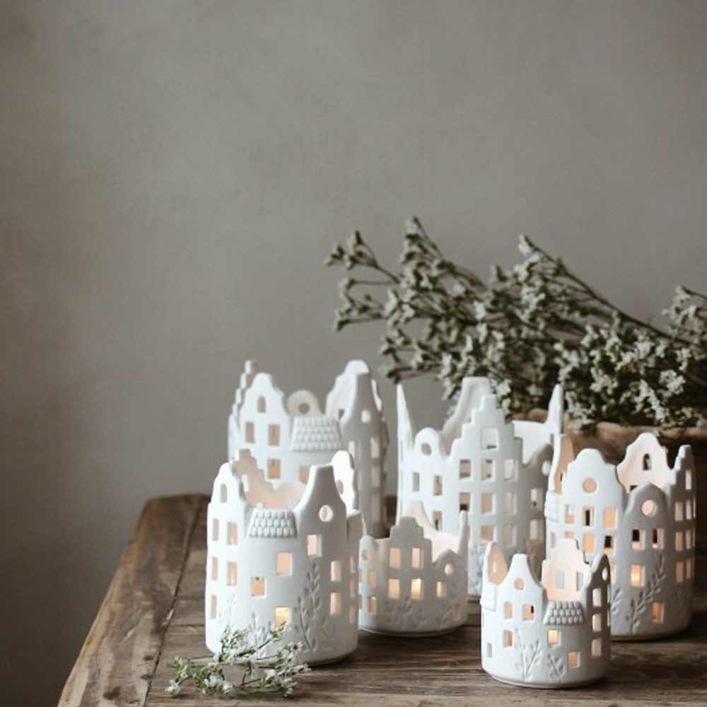 Majas Cottage – Lichthaus Keramik weißer Jardin in Form von kleinen Häusern mit brennenden Kerzen darin, auf einer Holzoberfläche neben einem Zweig getrockneter Blumen platziert.