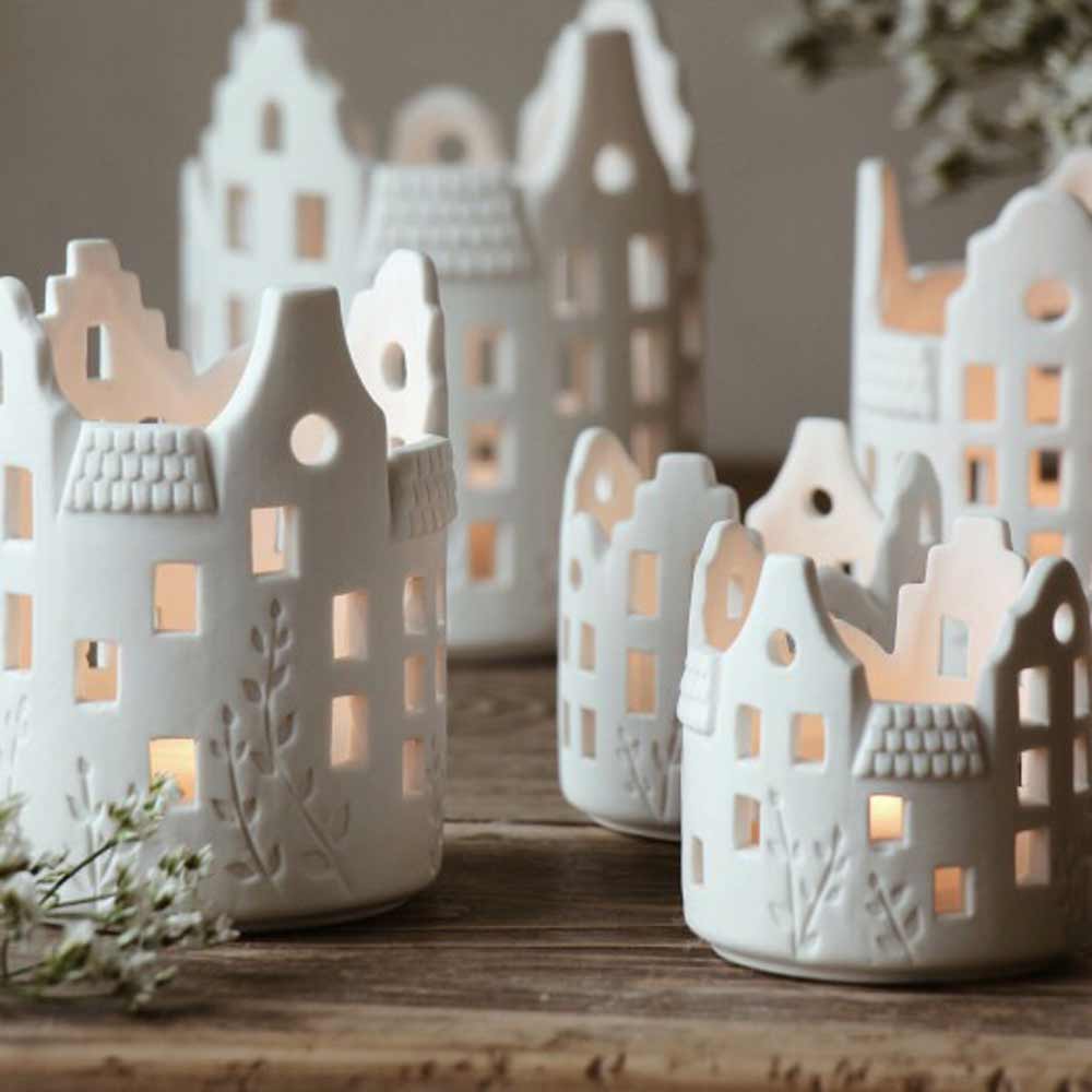 Eine Kollektion weißer Jardins von Majas Cottage – Lichthaus Keramik in Form kleiner Häuser mit von innen beleuchteten Fenstern, platziert auf einer Holzoberfläche mit Blumen im Hintergrund.