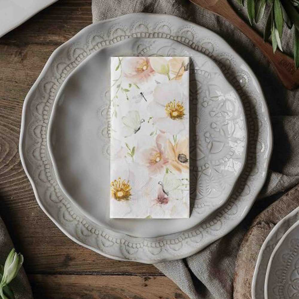 Die Majas Cottage Papierservietten Blumen & Schmetterlinge sitzen anmutig auf einem weißen Keramikteller mit kompliziertem Muster auf einem Holztisch, umgeben von frischem Grün.