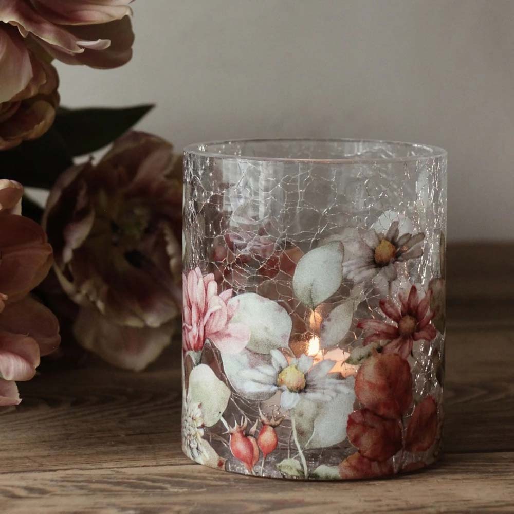 Eine brennende Kerze **Majas Cottage - Teelichthalter Blossom Bruchglasoptik Vintage** steht auf einer Holzoberfläche, umgeben von rosa Blumen.
