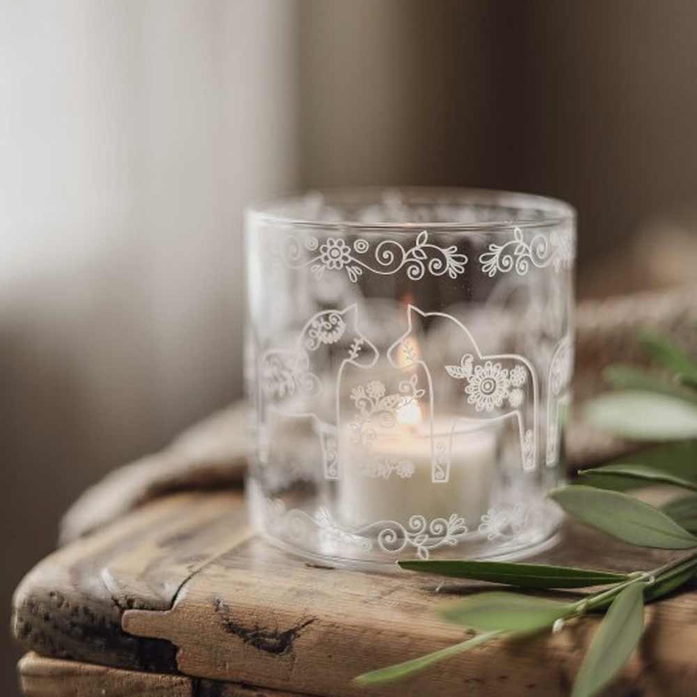 Der Majas Cottage Teelichthalter Dala Pferde Glas-Windlicht mit weißen Blumen- und Dalapferd-Motiven leuchtet mit einer brennenden Kerze auf einer rustikalen Holzoberfläche neben grünen Blättern.