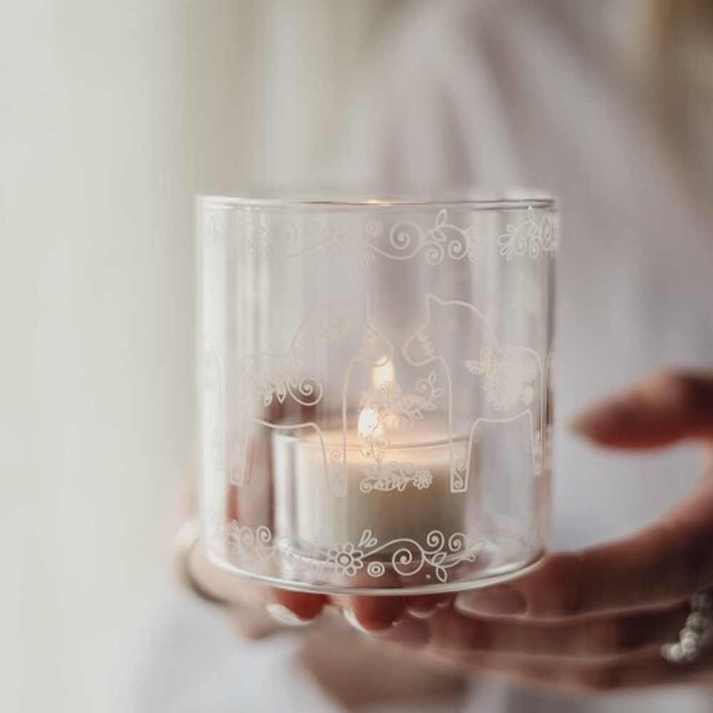 Jemand hält den Majas Cottage - Teelichthalter Dala Pferde Glas Windlicht in der Hand, einen Kerzenhalter aus klarem Glas mit weißen Blumenmustern und Pferdemotiven, in dem ein Teelicht leuchtet.