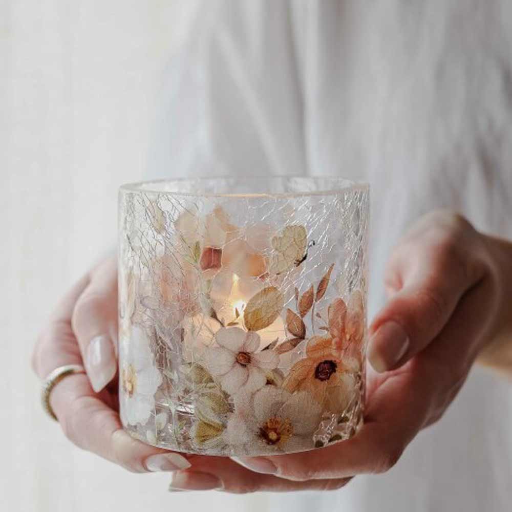 Eine Person hält den Glaskerzenhalter „Majas Cottage – Windlicht Blossom & Butterflies“, der mit gepressten Blumen und Schmetterlingen geschmückt ist und in dem sich eine brennende Kerze befindet.