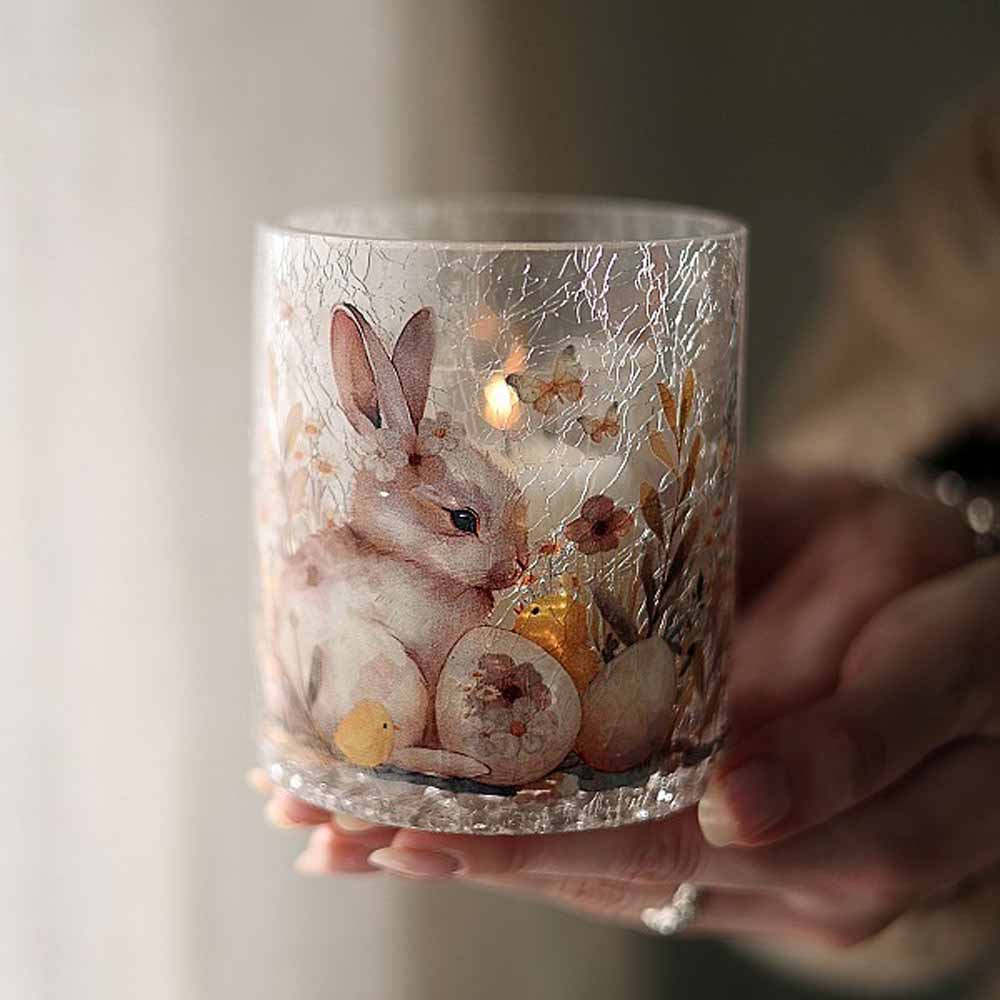 Eine Hand hält eine brennende Majas Cottage-Kerze „Windlicht Hase & Blumen“ in einem Glashalter, der eine Kaninchen- und Blumenillustration zeigt.