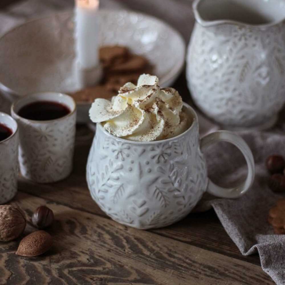 Der Majas Cottage - Winterheart Mugg ist mit Schlagsahne garniert und von Tassen mit dunkler Flüssigkeit, einer Kerze und verschiedenen Nüssen auf einem Holztisch umgeben.