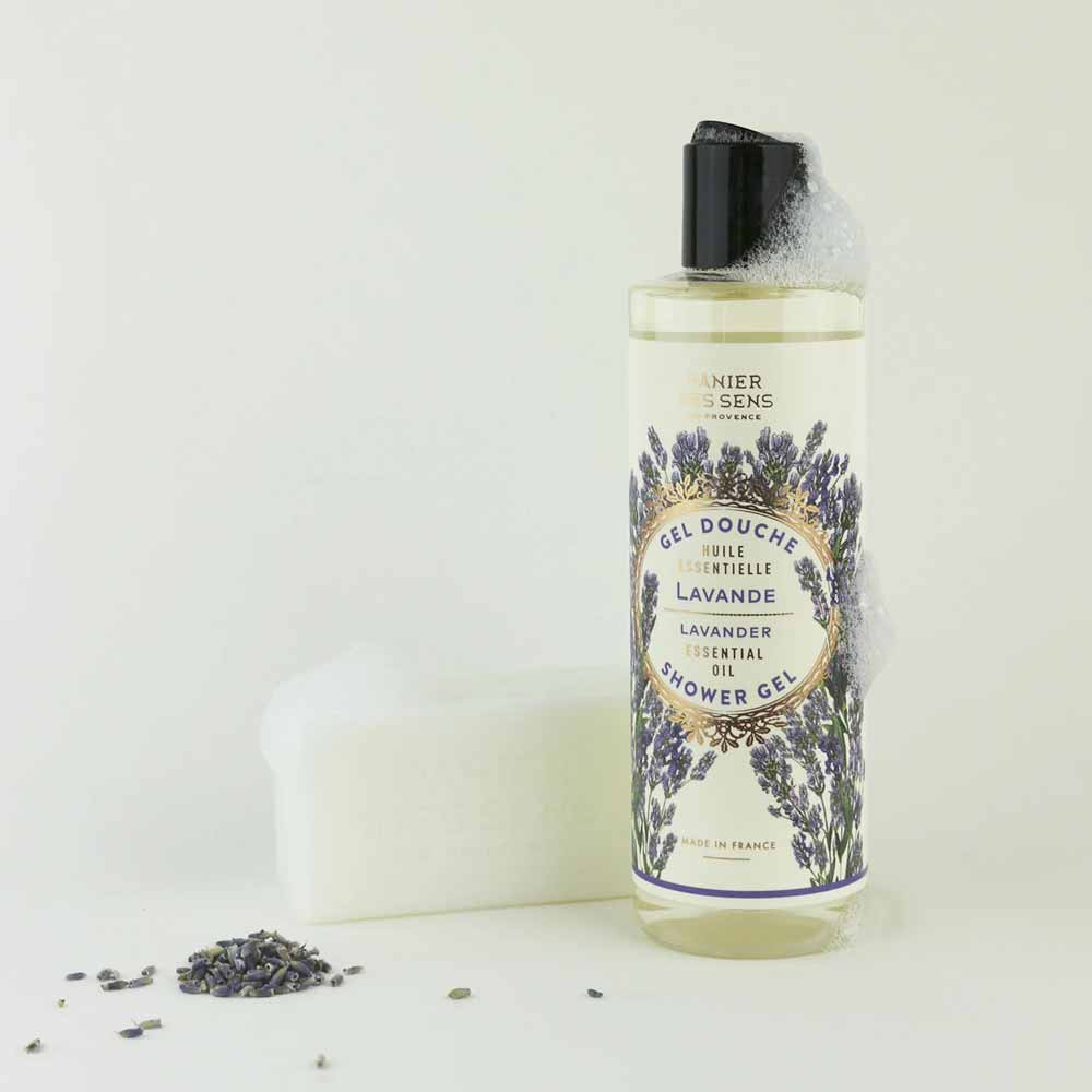Klare Flasche Panier des Sens – Duschgel Lavendel 250 ml mit Lavendelabbildungen auf dem Etikett, teilweise seifig, daneben ein weißes Stück Seife und einige verstreute Lavendelzweige.