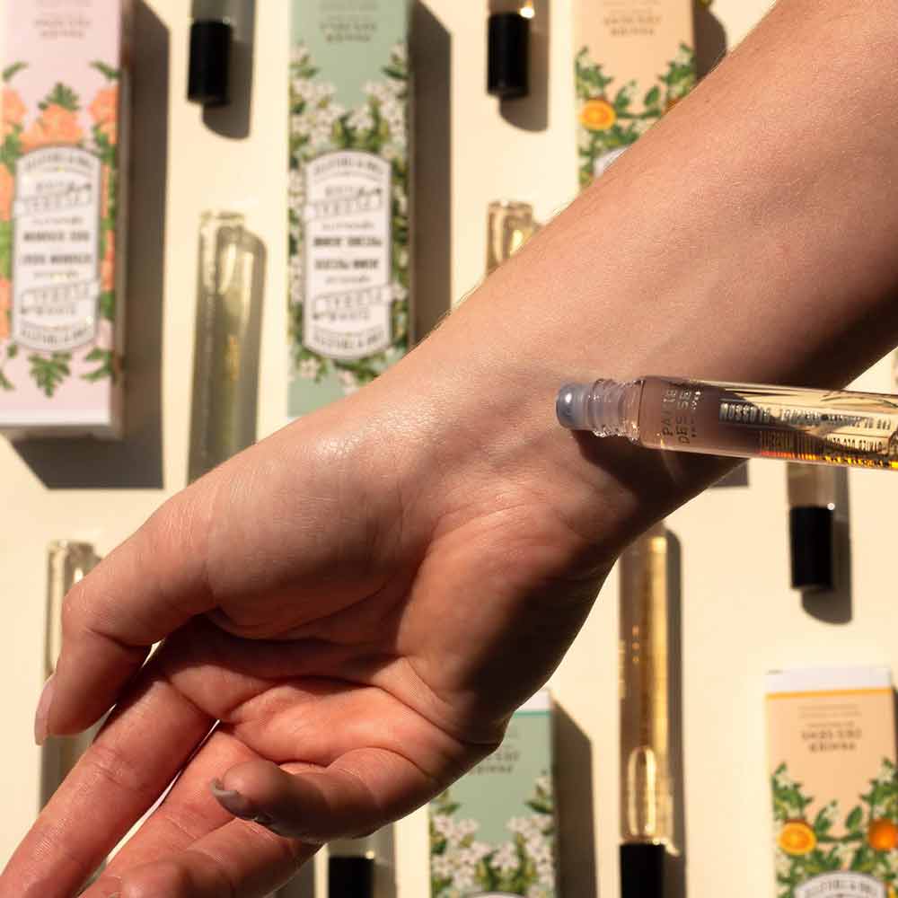 Eine Nahaufnahme einer Hand, die einen Panier des Sens – Eau de Toilette Roll-on Jasmin 10 ml auf das Handgelenk aufträgt, im Hintergrund sind verschiedene Parfümflaschen und Verpackungen zu sehen.