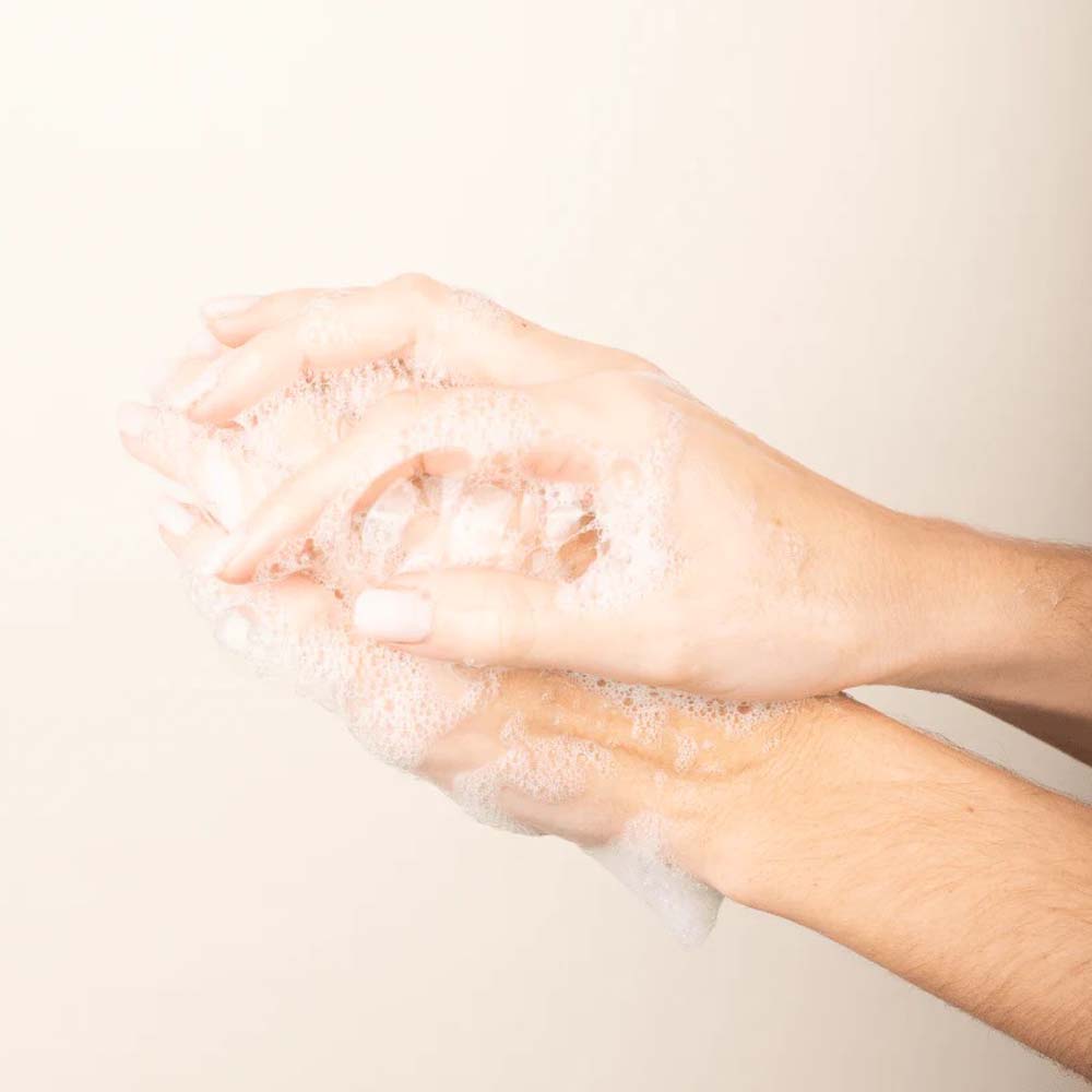Mit Seifenlauge „Panier des Sens – Feste Seife mit Duft Irisblütenblätter 150 g“ bedeckte Hände werden vor einem hellen Hintergrund gewaschen.