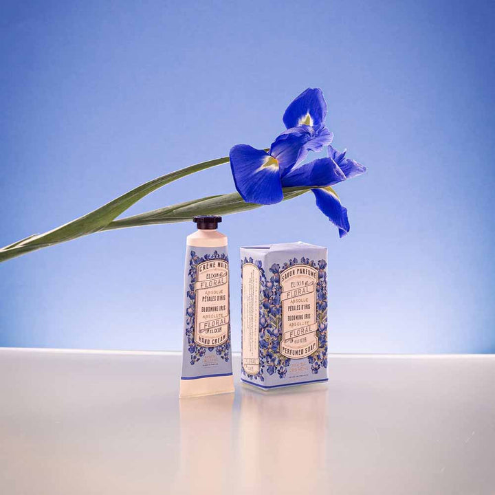 Eine einzelne Panier des Sens - Handcreme Irisblüttenblätter ruht auf zwei dekorativen Flaschen vor einem blauen Hintergrund mit Farbverlauf.