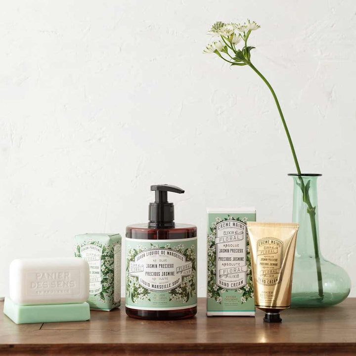 Eine Sammlung von Toilettenartikeln, darunter Flüssigseife, Seifenstücke und Panier des Sens – Handcreme Jasmin, ausgestellt neben einer grünen Glasvase mit einer einzelnen Blume.