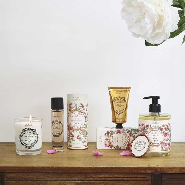 Eine Sammlung von Panier des Sens – Handcreme Rose, ordentlich präsentiert auf einer Holzoberfläche mit einer brennenden Kerze und weißer Blumendekoration.